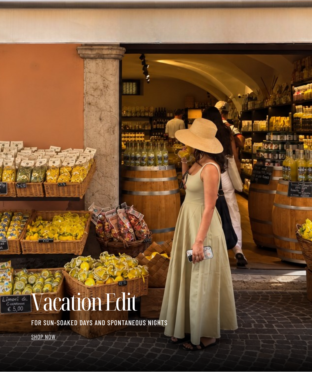 A woman in a long beige dress and wide-brimmed straw hat stands in front of a rustic shop doorway with barrels, shelves, and baskets of lemons and packaged goods. Large overlaid text reads 'Vacation Edit' with smaller copy below.