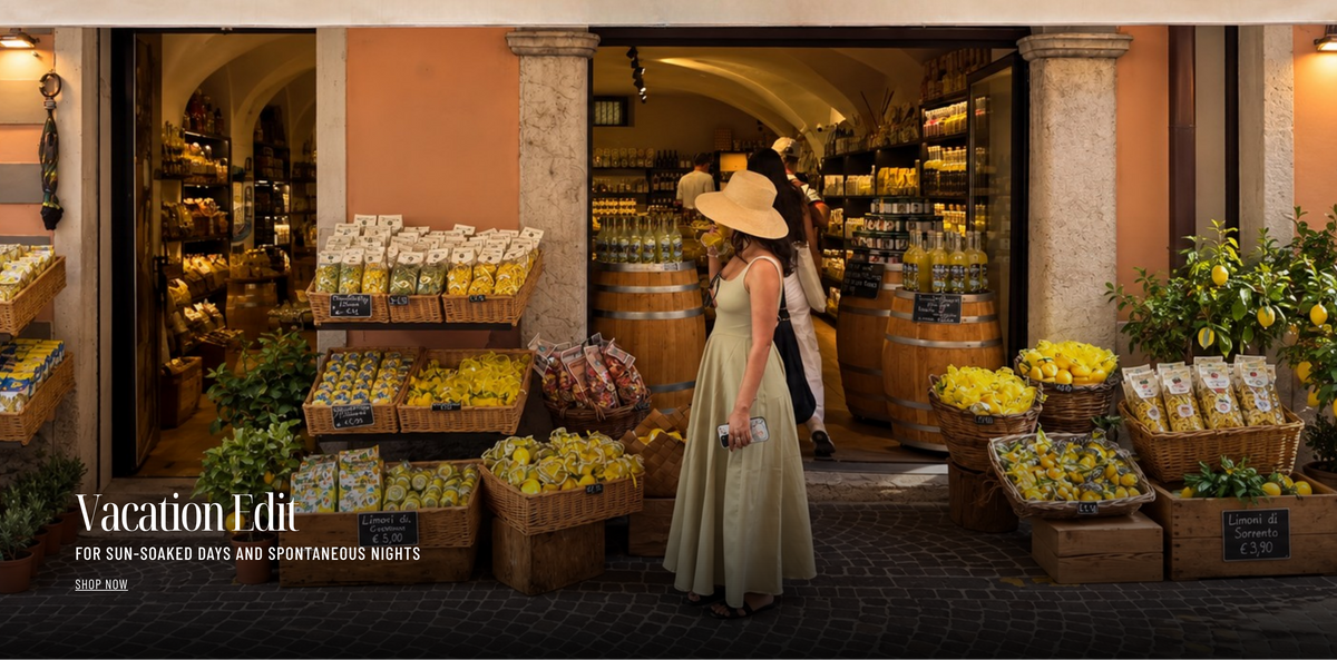 A woman in a long beige dress and wide-brimmed straw hat stands in front of an open storefront filled with bottled goods and surrounded by baskets of lemons and other produce. The scene is warmly lit and set in a real environment, with a visible 'Vacation Edit' text overlay in the lower left.